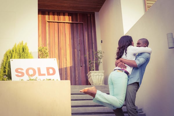 A woman and a man are hugging on the steps of a home. With a beautiful bamboo door and potted tree out front. A SOLD sign is placed in the foreground.