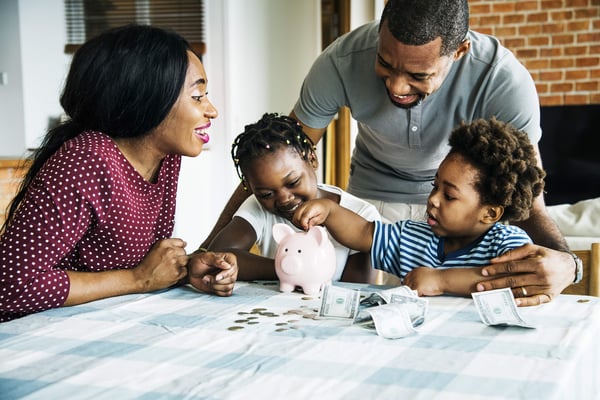 A family of four happily gather around a table where their young children place coins and dollar bills into a small pink piggy bank.