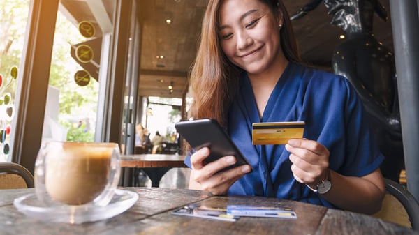 A well dressed woman smiles as she sits at a table with a cell phone in his right hand, and a credit or debit card in his left hand.